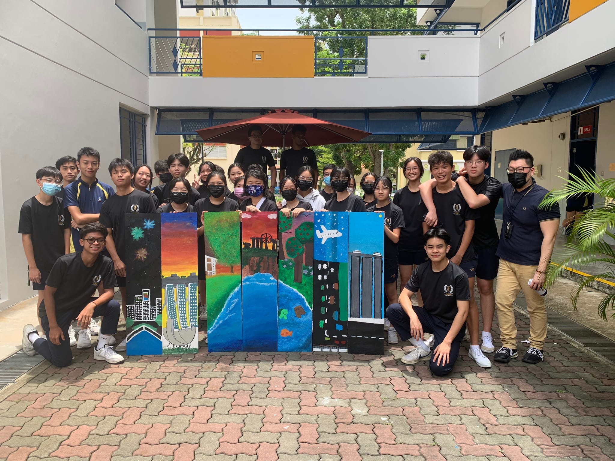 A group of students in black uniforms pose outdoors with colorful painted panels showing cityscapes and nature scenes.